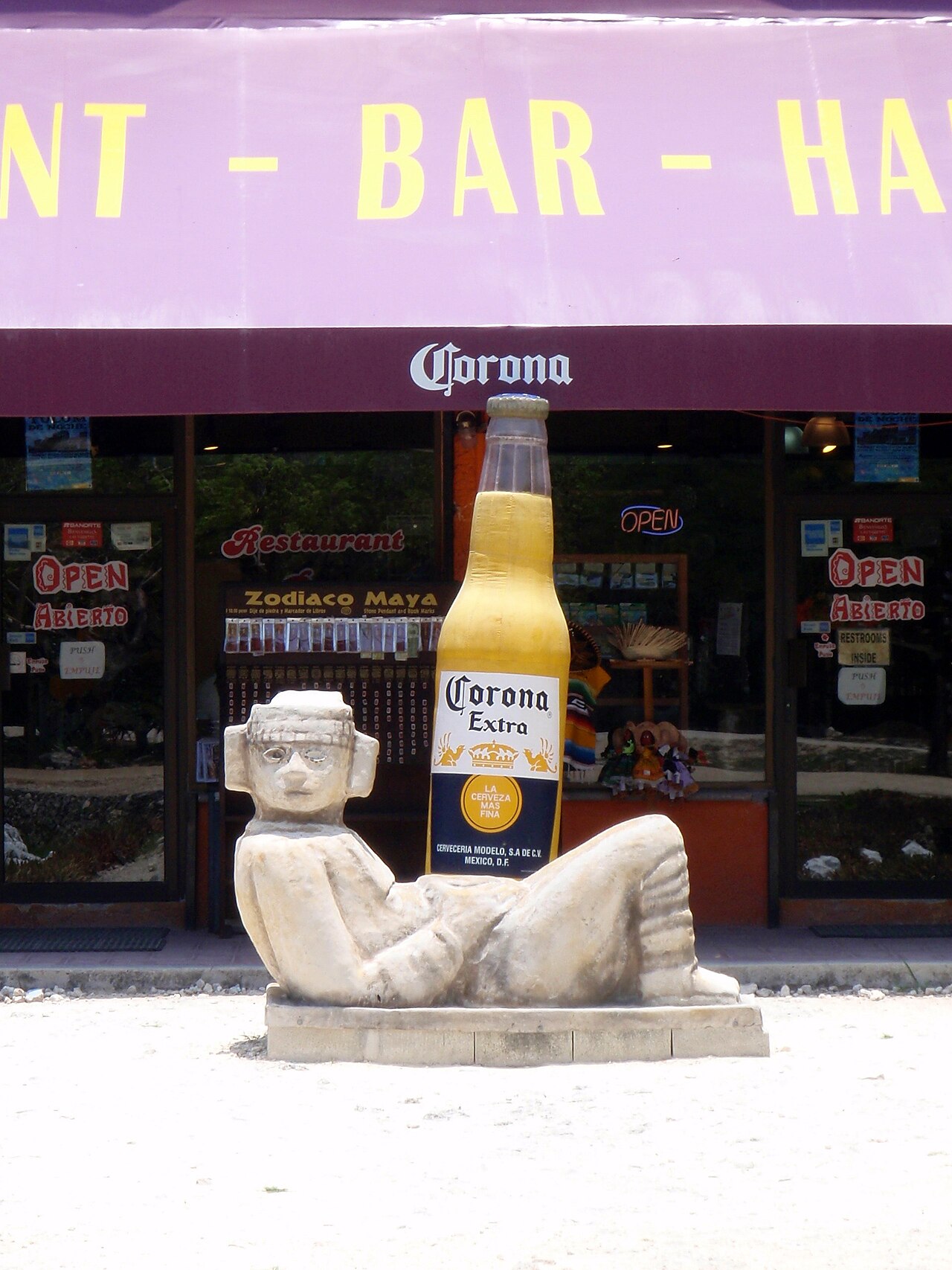 A bottle of Corona Extra standing beside a stone Chac Mool figure under the sun in Tulum, Mexico