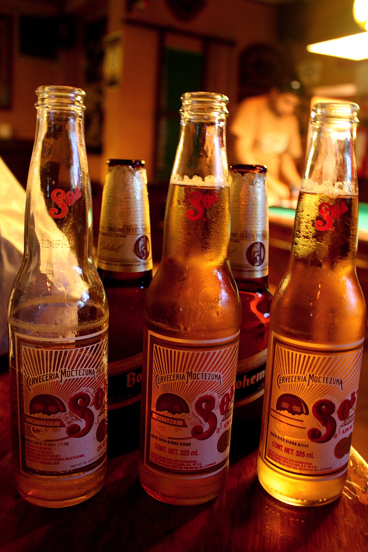 Three bottles of Sol cerveza with their Cervecería Moctezuma sunburst labels on a wooden bar table, pool table glowing behind