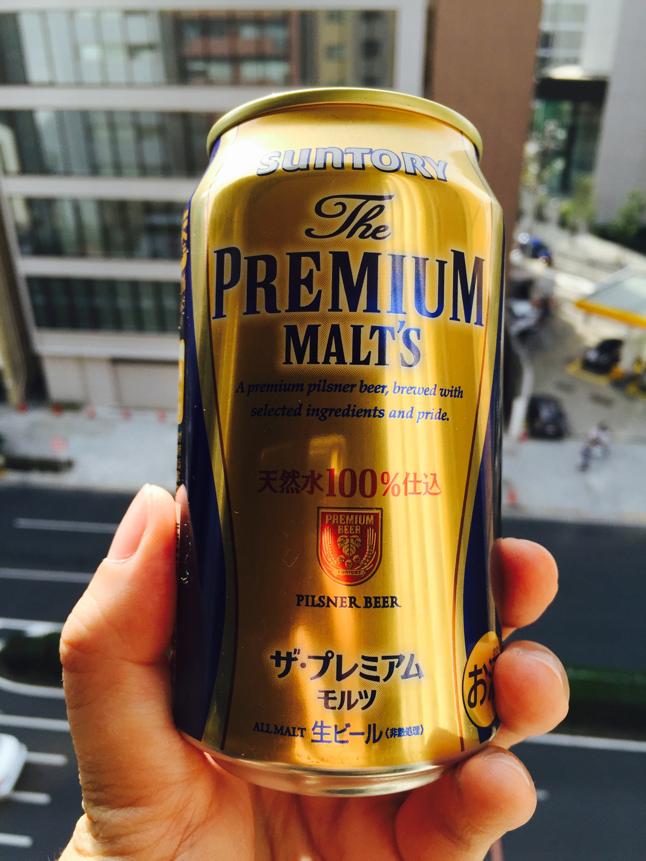Hand holding a gold Suntory The Premium Malt's can above a sunlit Japanese street